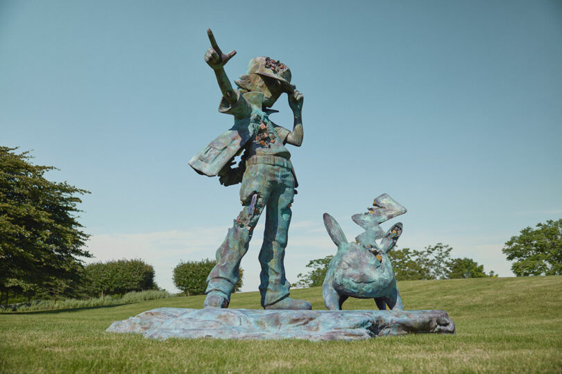 A bronze statue of a person in a hat holding a device with one hand and raising the other, standing next to an animal with large ears on a grassy field. Trees and a clear sky are in the background.