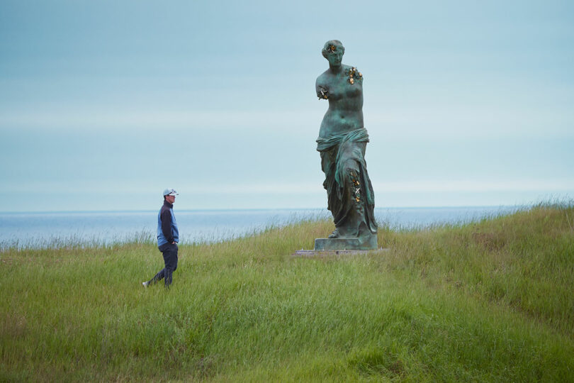 A person wearing a hat walks near a large green statue of Venus de Milo situated in a grassy landscape with the sea in the background.