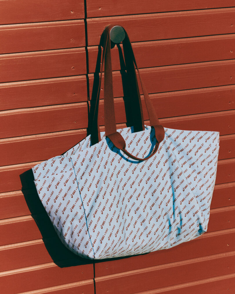 A blue tote bag with brown leather straps, featuring a repeating red text pattern, hangs on a circular hook against a red slatted wall, perfectly complementing the style of ASICS x HAY collaboration.