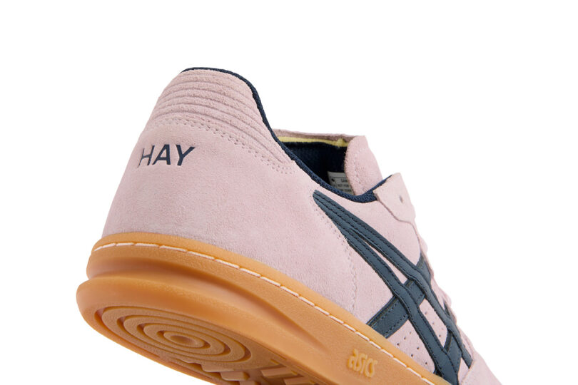 Close-up of a light pink ASICS x HAY sneaker with a gum sole, featuring dark blue stripes and the word "HAY" embroidered on the heel. The shoe rests on its side, showcasing the intricate sole pattern.