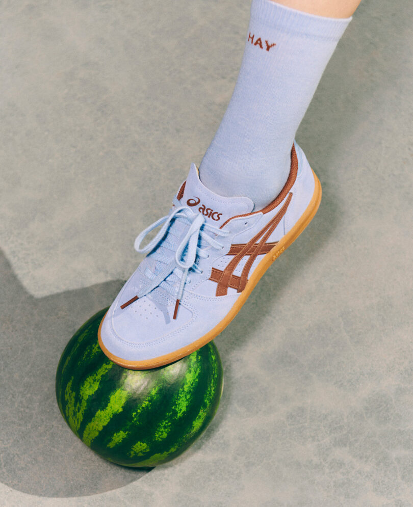 A foot wearing a blue and brown ASICS x HAY sneaker and a light blue sock stands on a watermelon.