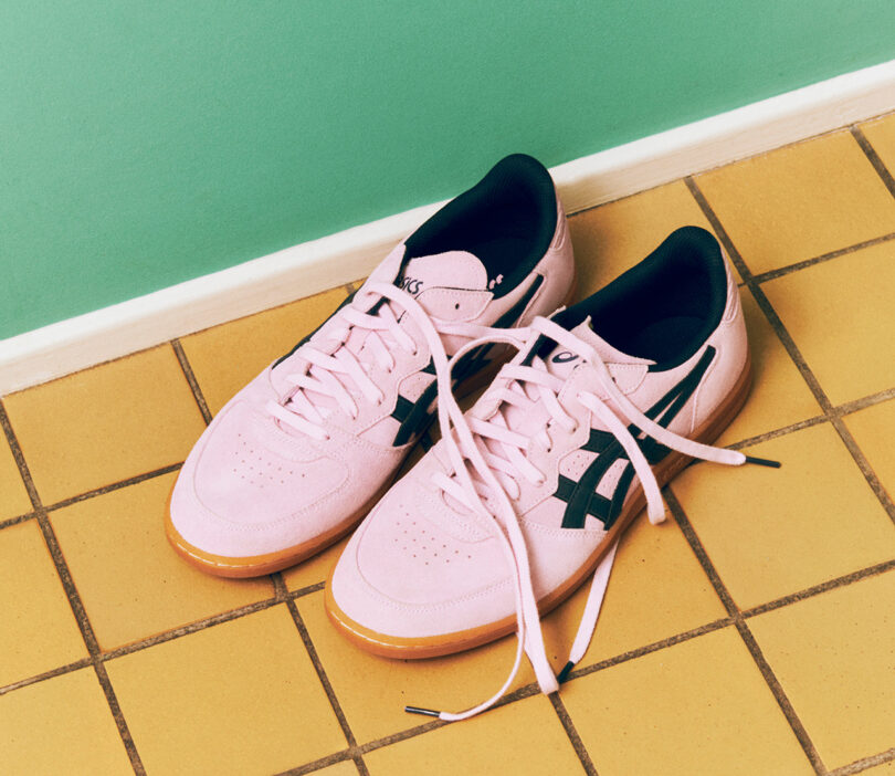 A pair of pink ASICS x HAY sneakers with black accents and untied laces sits on a tiled floor next to a green wall.