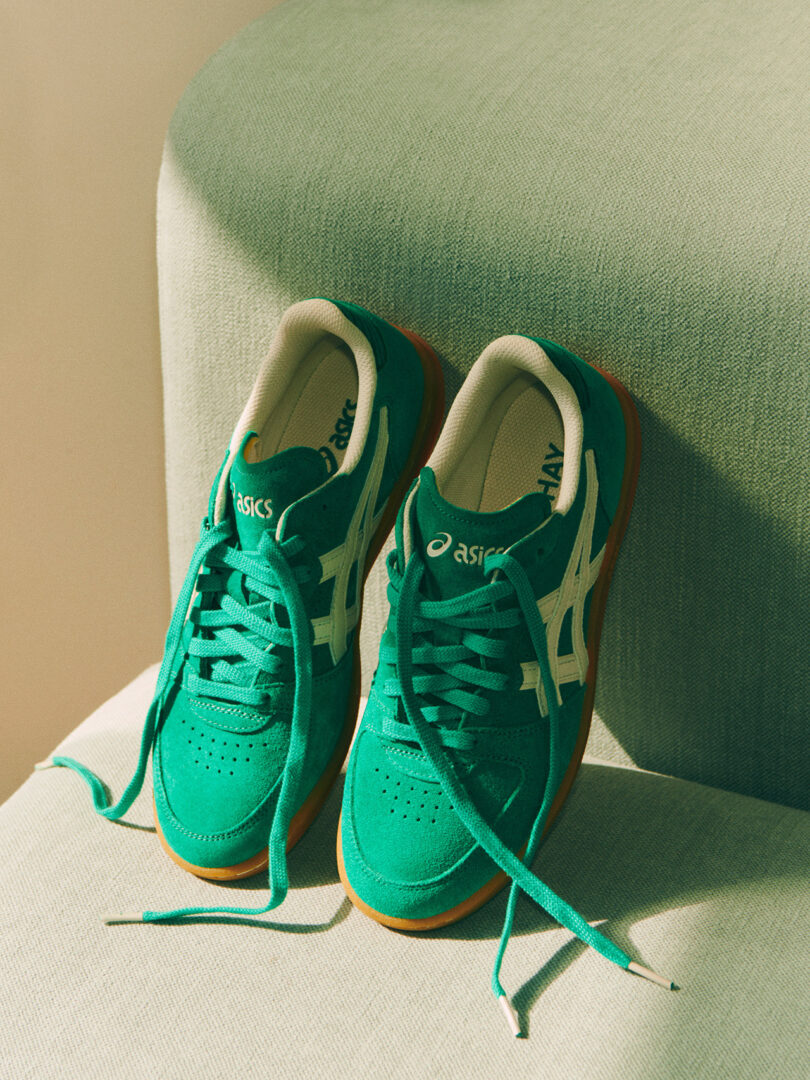 A pair of green and white ASICS x HAY sneakers with untied laces, resting on a beige fabric surface.