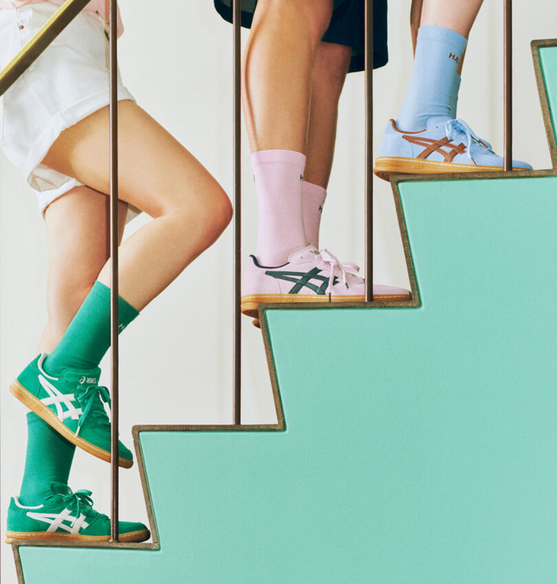 Three people standing on stairs, wearing colorful sneakers and socks. One person sports green sneakers and socks, another flaunts pink ASICS x HAY sneakers with blue socks, and the third pairs beige sneakers with blue socks.
