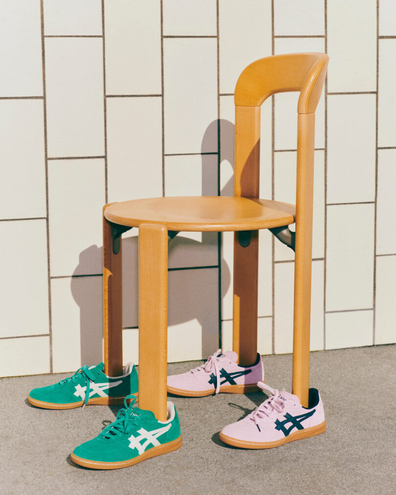 A wooden chair with three pairs of ASICS x HAY sneakers positioned under it, resembling legs. The pairs include green, pink, and blue sneakers with a white logo, placed on a tiled floor against a white tiled wall.