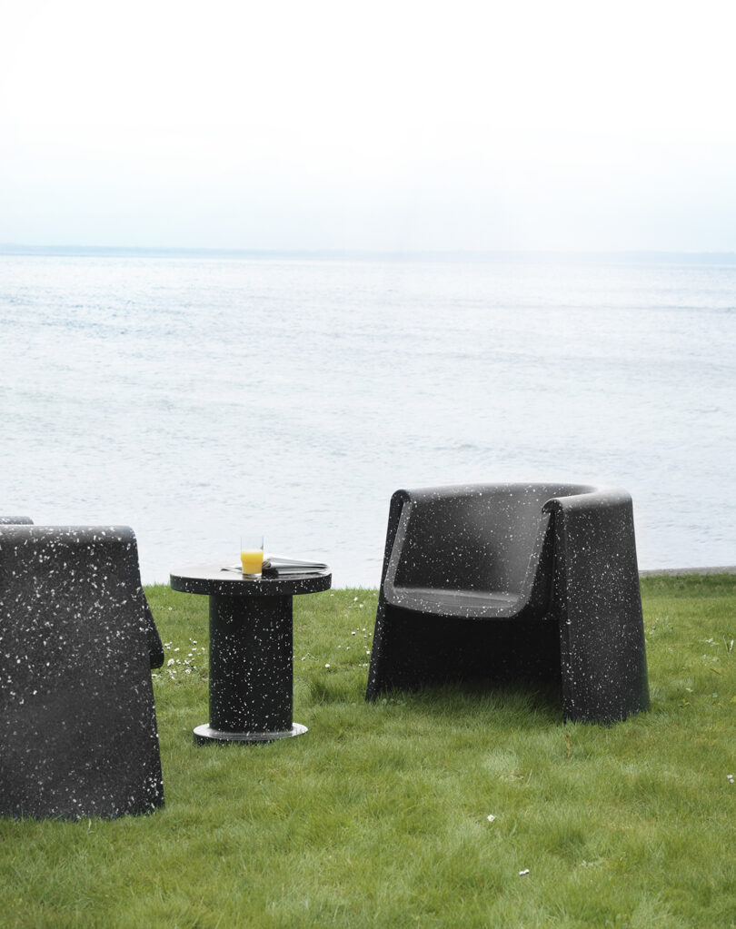 Two black speckled lounge chairs and a matching table with a glass of orange juice are set on a grassy area near a body of water under a cloudy sky.