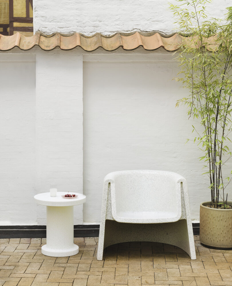 A white lounge chair and round table are positioned against a white brick wall with a clay tile roof. The table holds a glass and a small dish. A bamboo plant in a pot stands to the right.