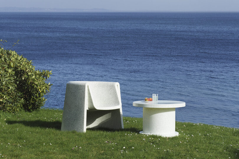A white lounge chair and round table are placed on a grassy area near a body of water. The table has a glass of water and a plate with fruit.