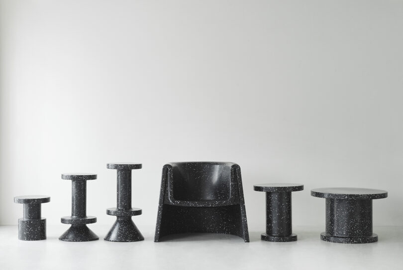 A collection of black speckled minimalist furniture, including stools, tables, and an armchair, arranged in a row against a plain white wall.
