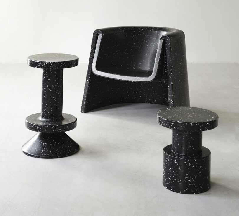 A black, speckled armchair is flanked by two matching, uniquely shaped side tables on a plain, light-colored floor.