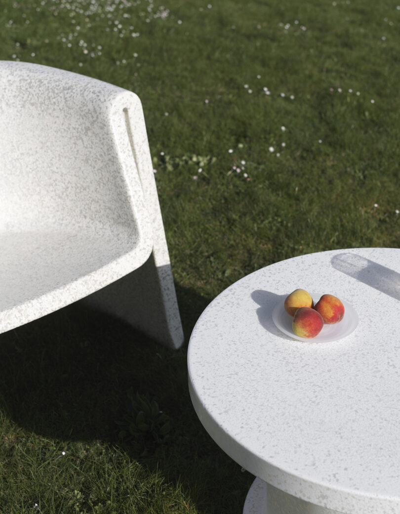 A white chair and round white table are on a grassy lawn. The table has a plate with three peaches on it.