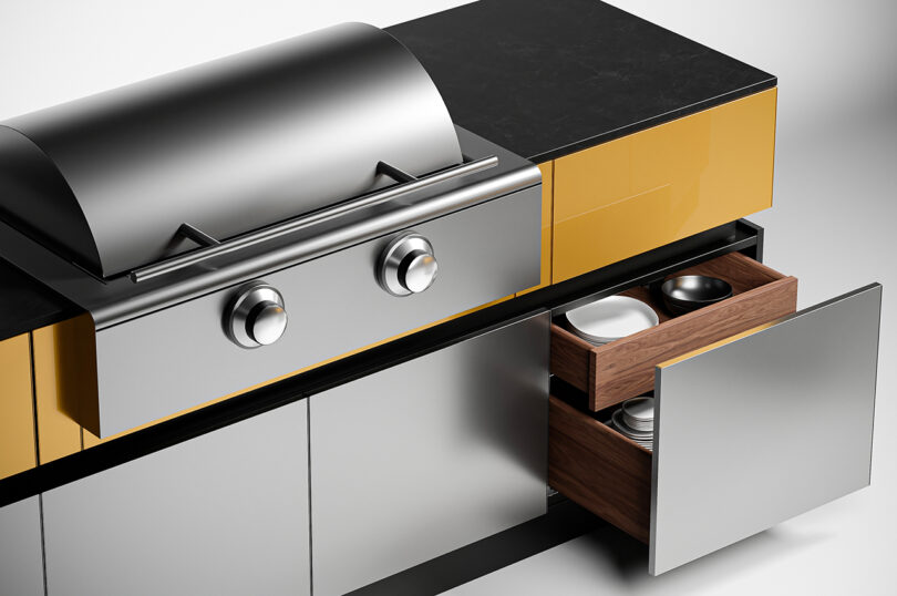 A stainless steel outdoor kitchen and grill with two control knobs and a closed lid, next to a yellow and grey storage unit with an open drawer and cabinet containing dishes and bowls.