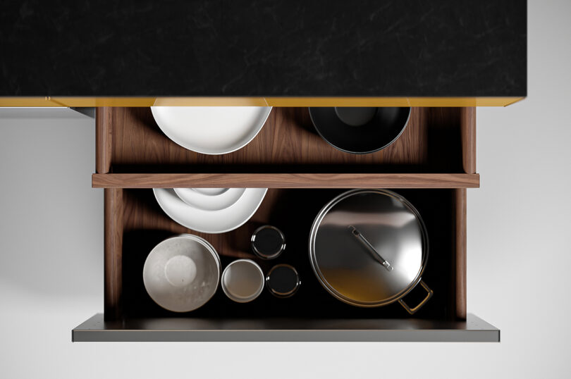 Top view of an open kitchen cabinet drawer containing neatly arranged plates, bowls, cups, and a large stainless steel pot with lid. The interior of the drawer is wooden.