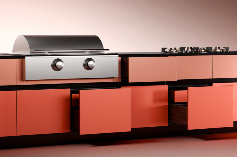 A modern outdoor kitchen with coral colored cabinets, open drawers, a stainless steel oven with two knobs, and a gas cooktop with three burners.