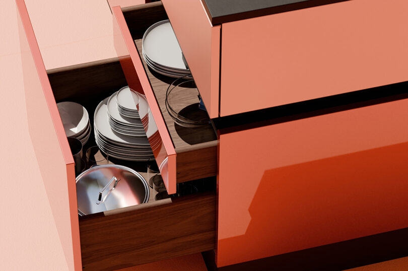 Open kitchen drawers filled with neatly stacked white plates, bowls, and cookware. The cabinetry is a glossy coral color.