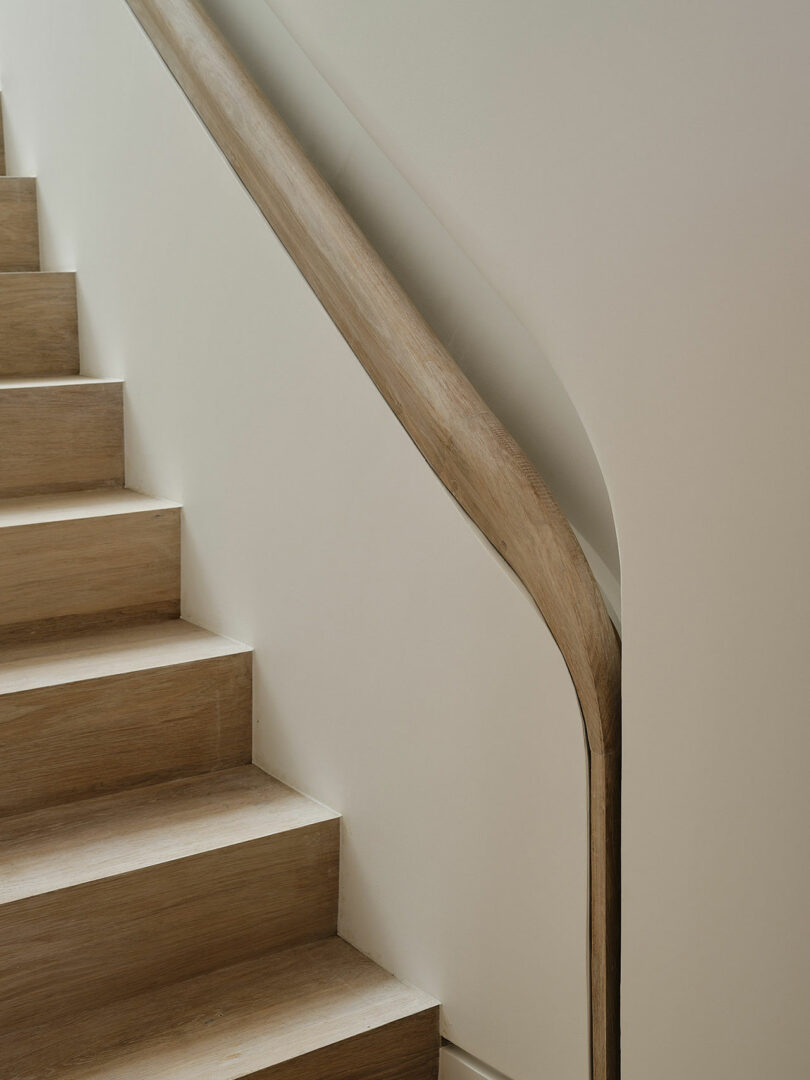 A wooden staircase with light brown steps and a matching handrail against a white wall. The handrail follows the curve of the stairway.