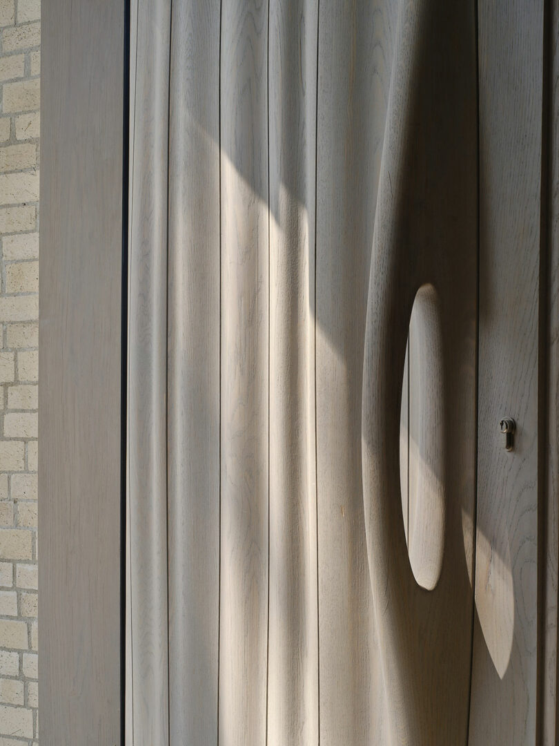 A close-up of a modern wooden door with a unique curved handle design, partially illuminated by sunlight. The adjacent brick wall is visible on the left side.