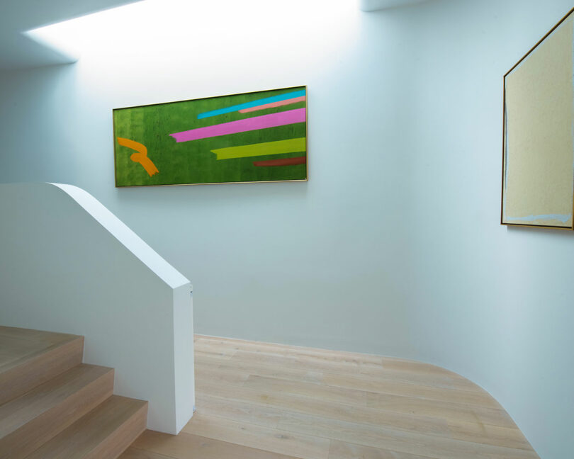 A minimalist interior with white walls, wooden floors, and a staircase. Colorful abstract painting hangs on the right wall, and another neutral-toned painting is partially visible on the left.