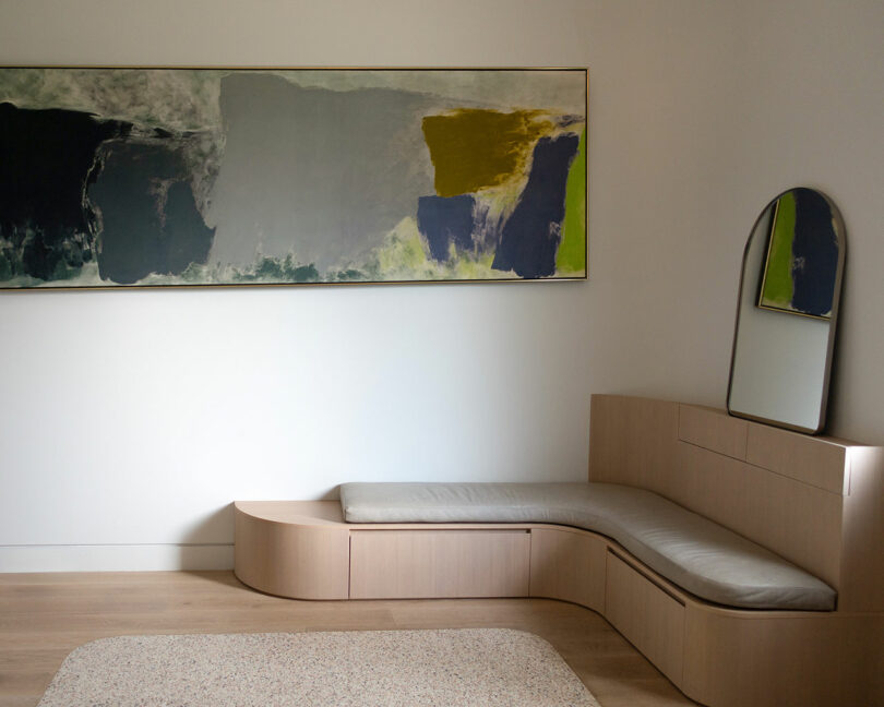 A minimalist room features an abstract painting on the wall, a beige cushioned bench with a built-in mirror on the right, and a neutral-colored rug on a wooden floor.