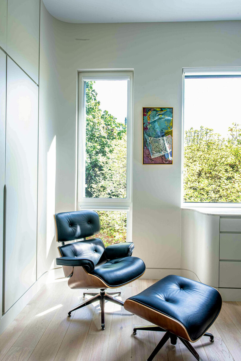 A modern room with a black leather lounge chair and matching ottoman by a window, with tall white walls and a piece of abstract art hanging above.