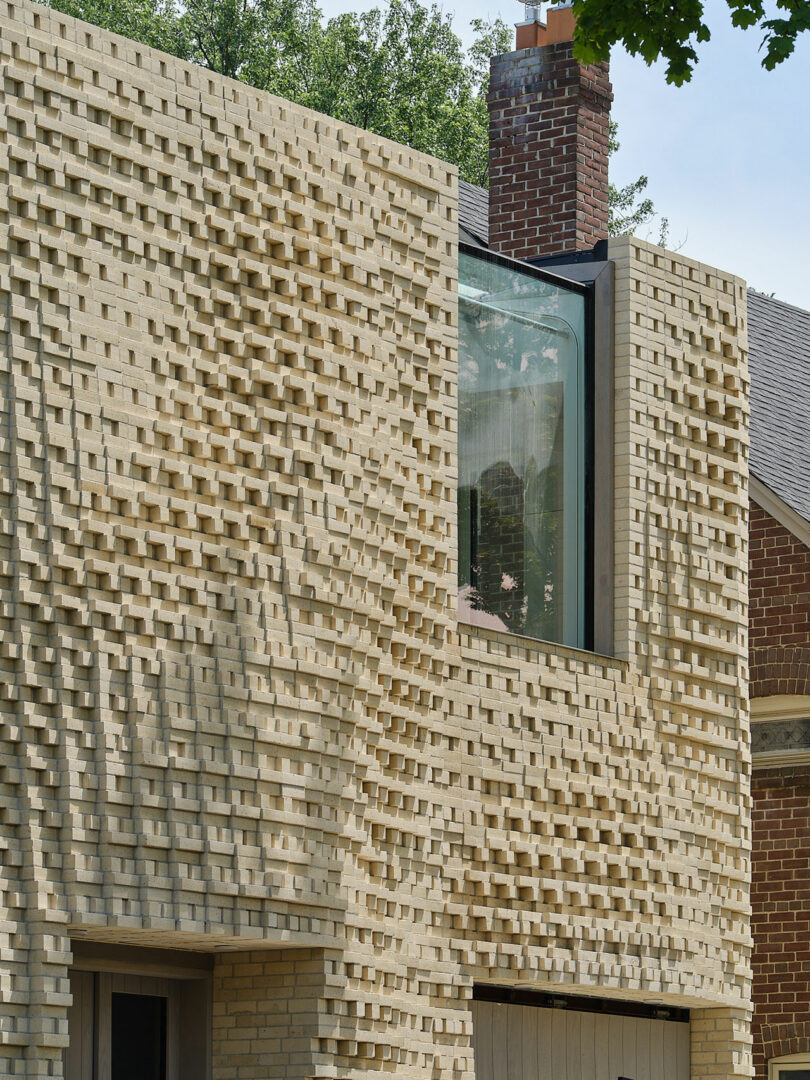 A building facade showcasing a textured, brick pattern with an inset window near the center. The upper part of the facade features a slight overhang. The background includes trees and part of a slanted roof.