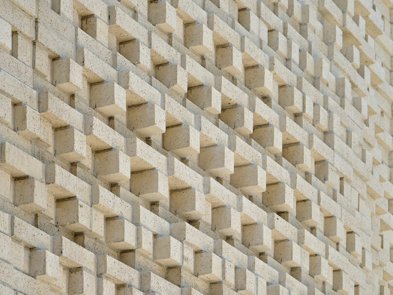 Close-up of a beige brick wall featuring a repetitive pattern of protruding and recessed rectangular blocks, casting small shadows and creating a textural, geometric design.