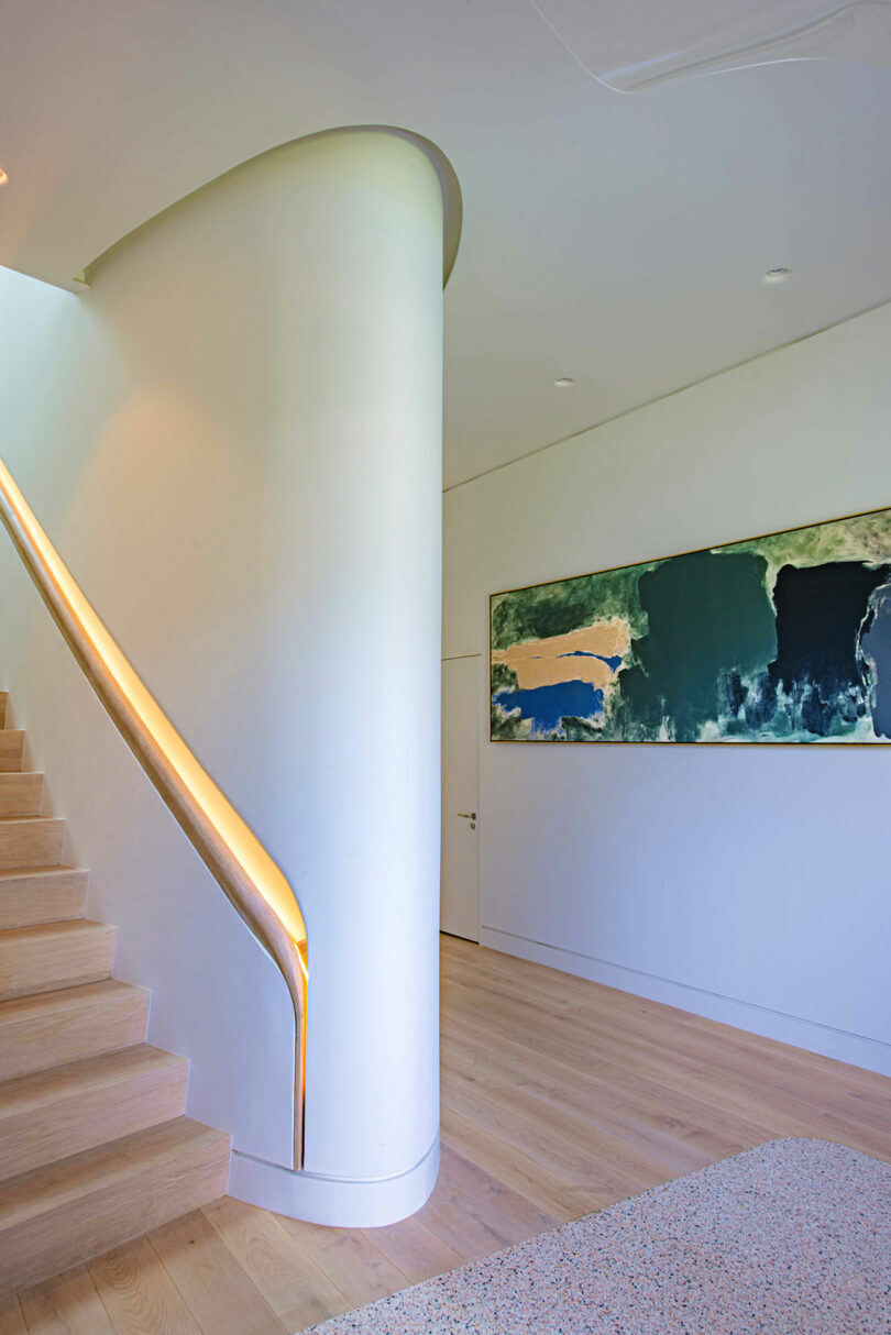 A modern interior featuring a curved staircase with a lit handrail, light wood steps, and a large abstract painting on a white wall.