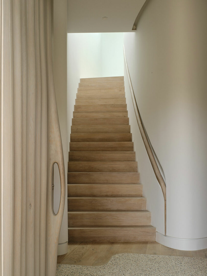 Minimalist wooden staircase with a curved handrail leading upstairs, framed by smooth walls and light wooden accents. A section of a curvilinear wooden structure is visible on the left side.