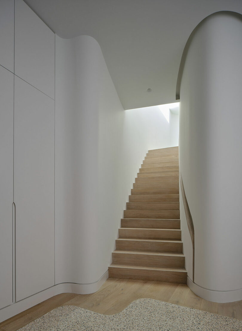 A minimalist interior featuring a wooden staircase with a sleek, curved white wall, under a skylight. The adjacent wall has built-in cabinets with smooth, handleless doors.
