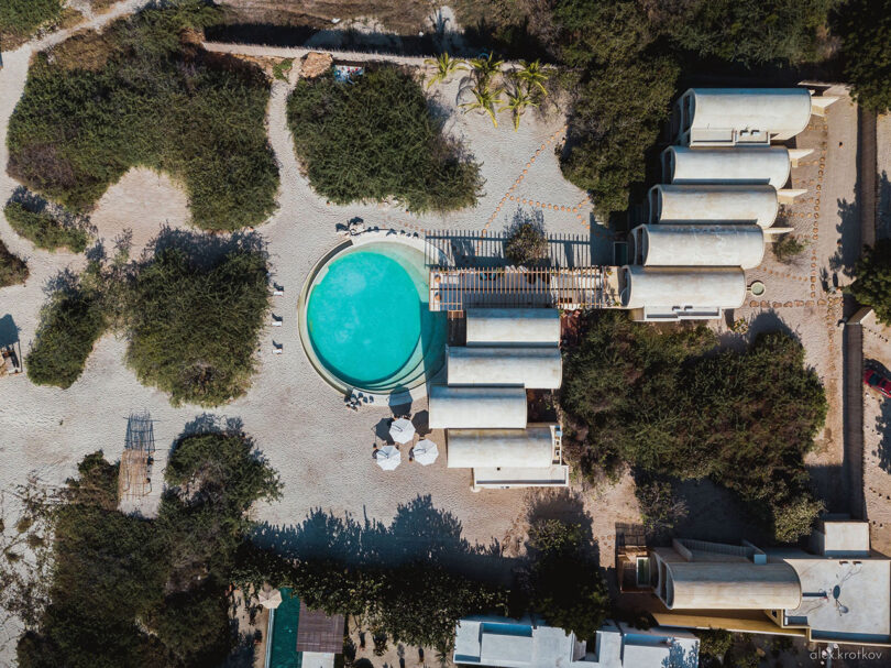 Aerial view of Casona Sforza in Puerto Escondido with a round salt water swimming pool surrounded by modern cylindrical buildings and sparse vegetation, set in a sandy, open area. The scene has a serene, minimalist aesthetic reminiscent of Casona Sforza in Puerto Escondido, Oaxaca Mexico.