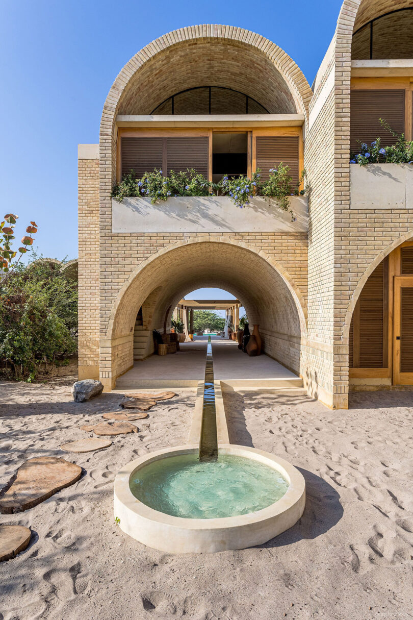 A brick building with arched doorways and open balconies features a narrow water feature extending from a circular basin, reminiscent of the architectural charm found in Oaxaca, Mexico.