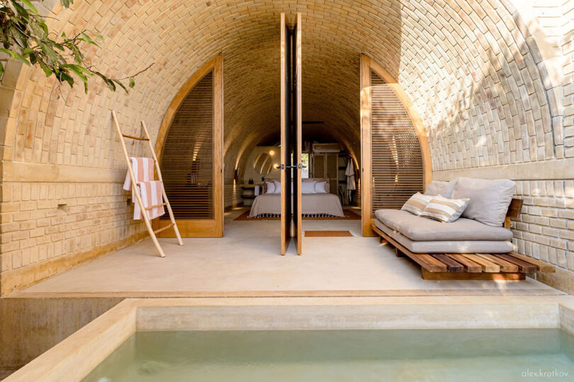 An indoor bedroom with open wooden doors leads to a patio with a cushioned bench, overlooking a small pool. The arched ceiling and walls, made of light brick, evoke the charm of Casona Sforza in Puerto Escondido, Oaxaca Mexico. A striped towel hangs on the left.