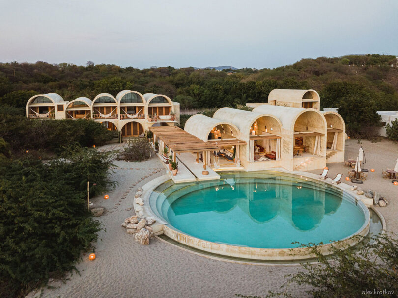 Aerial view of Casona Sforza in Puerto Escondido, Oaxaca, Mexico. This luxurious beach resort boasts arched architecture and features a large circular pool surrounded by loungers and greenery, set against sandy terrain and lush foliage.