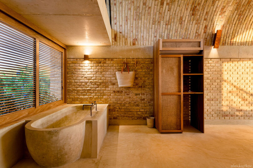 Modern bathroom with a stone bathtub, wooden blinds, brick walls, and warm lighting inspired by the aesthetics of Casona Sforza in Puerto Escondido. A wooden cabinet with open shelving is on the right side of the room, evoking the rustic charm of Oaxaca Mexico.