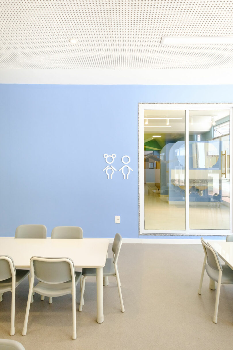 A minimalist room with light blue walls, white furniture, and a set of four chairs around a table. A glass door separates this room from another space. Simple wall icons indicate gender-neutral restroom.