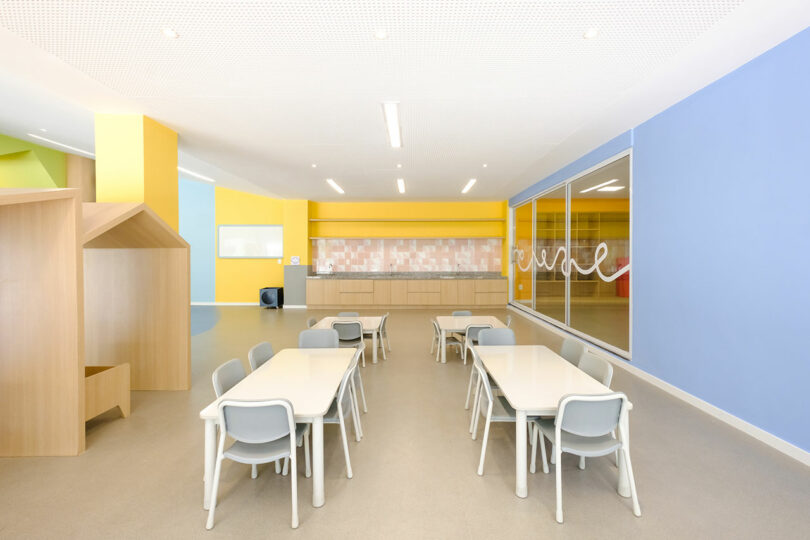 A brightly lit classroom with light blue chairs and white tables, wooden furniture, colorful walls, large windows, and a kitchenette area at the back.