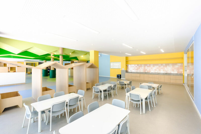 A brightly lit, modern kindergarten classroom with white tables, gray chairs, colorful walls, and wooden play structures resembling small houses.