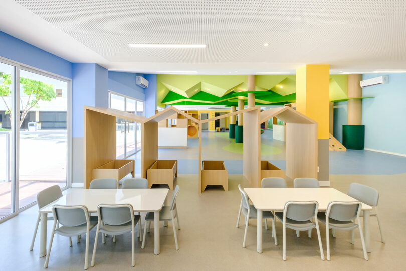 A bright, spacious room with tables and chairs, featuring wooden house-shaped partitions and colorful walls, designed for a children's play or learning area. Large windows allow natural light to enter.