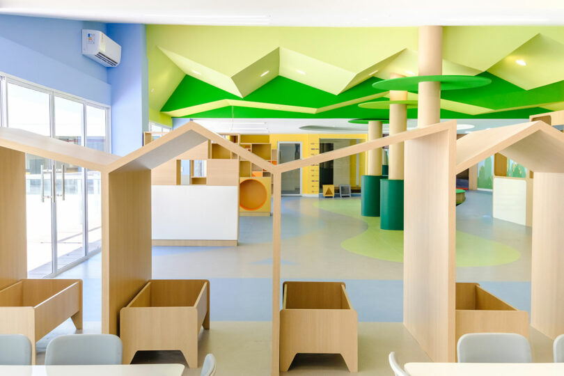 A brightly lit children's play area with geometric green ceiling panels, light wood play houses, and colorful furniture. Large windows allow natural light to flood the space.