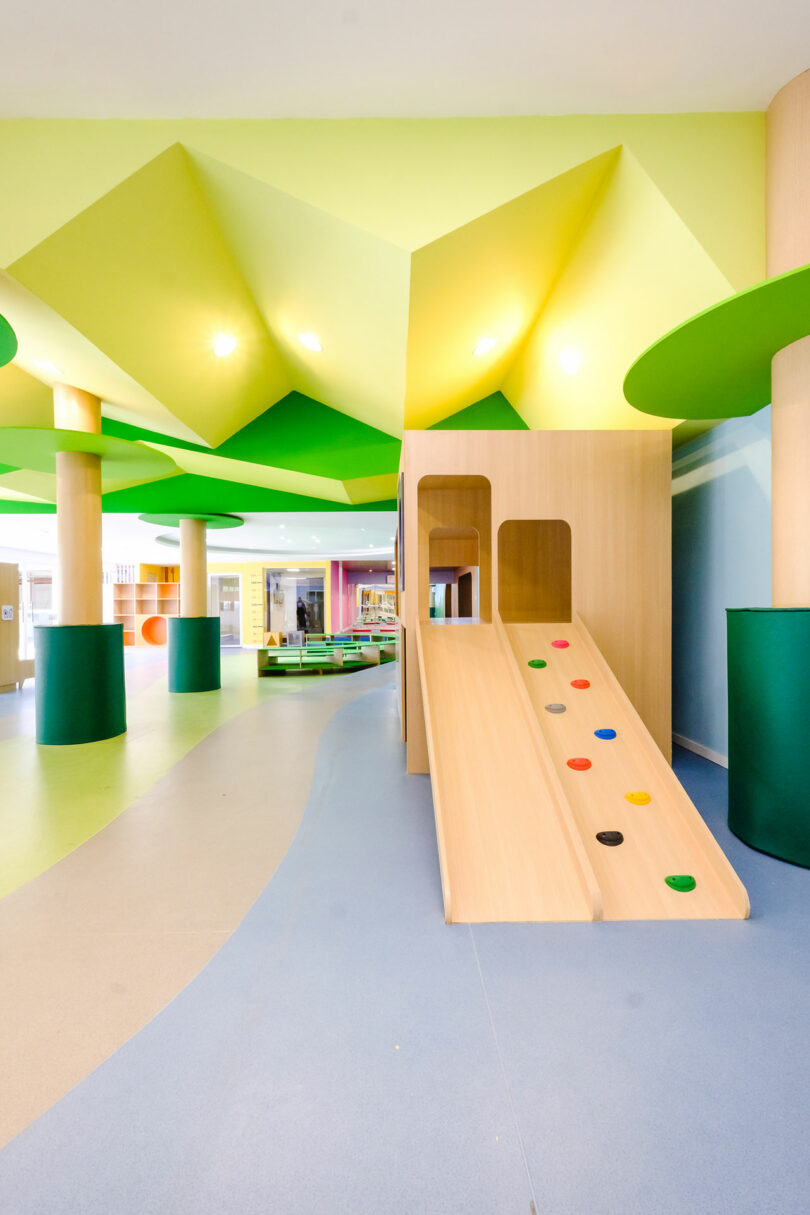 A brightly lit indoor playground with a wooden slide, colorful climbing knobs, and green column accents. The floor is a mix of blue and green tones, and there is open space for children to play.