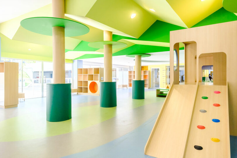 An indoor play area with green and yellow ceiling, wooden columns, shelving units, and a slide with colorful climbing grips.