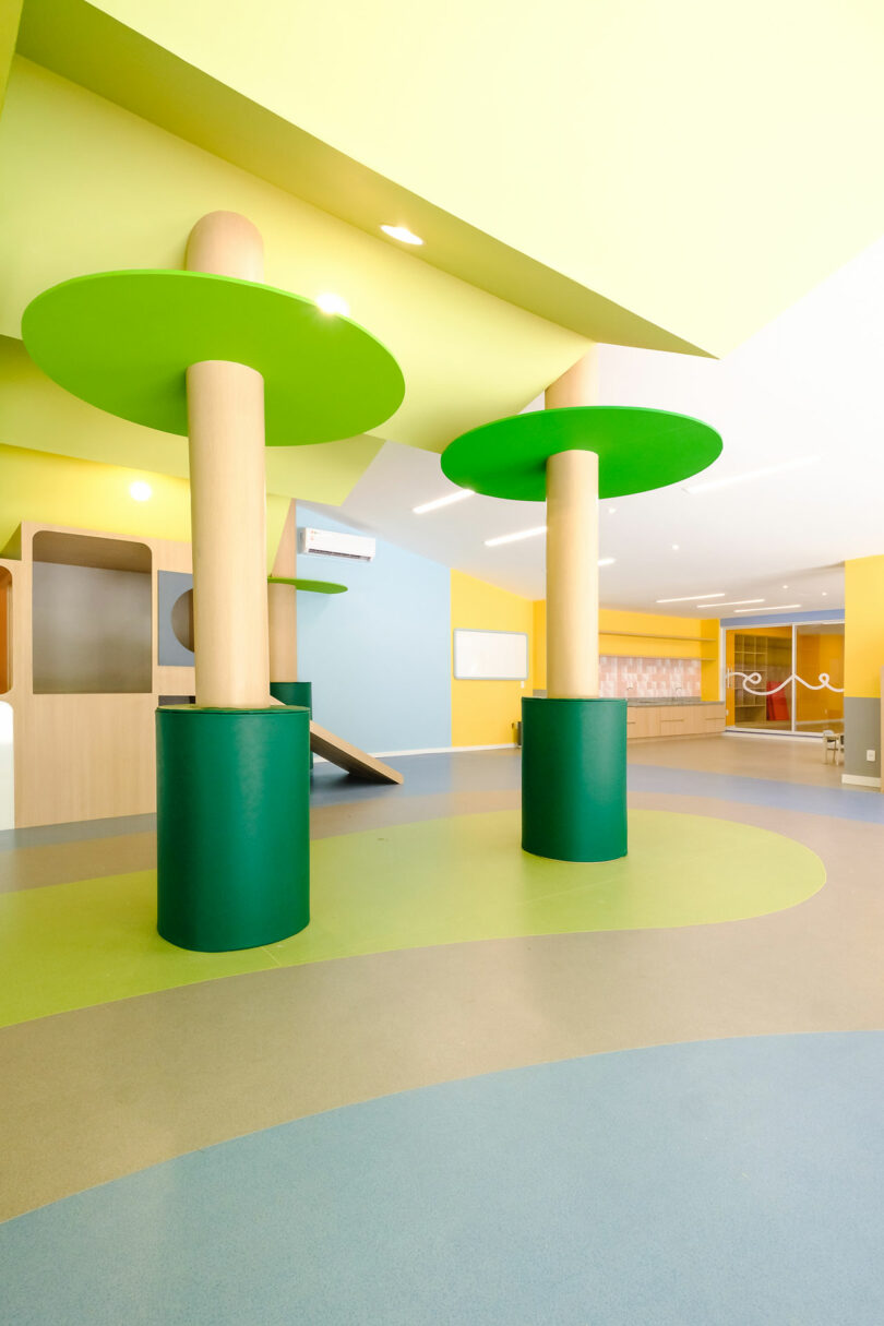 A brightly lit room with colorful decor, featuring large cylindrical columns resembling trees, soft flooring in shades of blue and green, and playful structures for children.