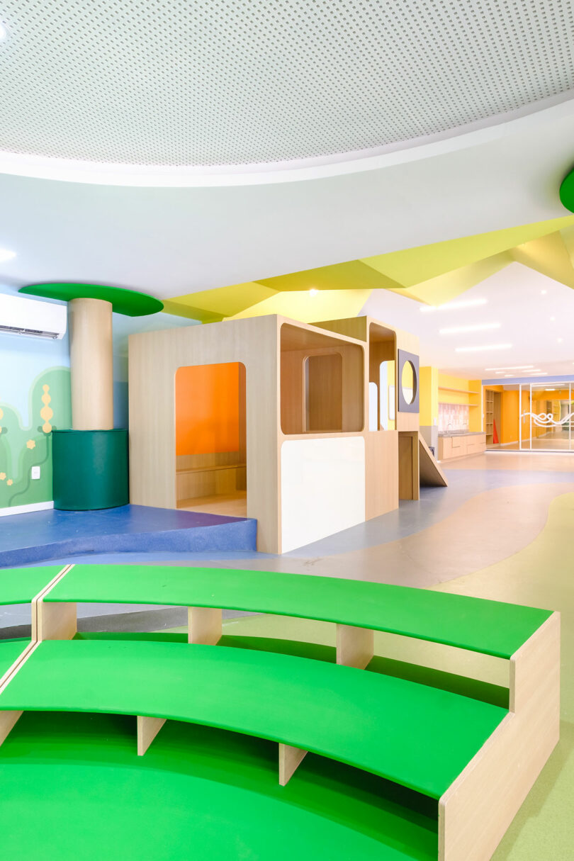 A brightly colored indoor playground with green seating, a playhouse structure, and vibrant walls. The space has a colorful and playful design, with various play elements for children.