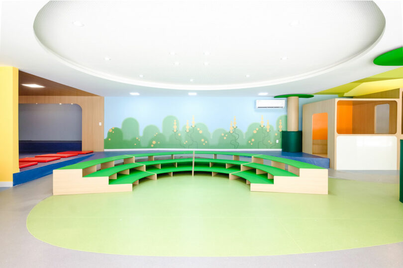 A colorful classroom featuring a semi-circular wooden seating arrangement with a green carpet and a mural of a sunny landscape on the wall.