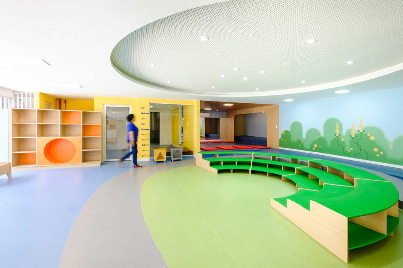 Bright, spacious children's play area with green circular seating, colorful wall murals, open shelves, and a walking individual.