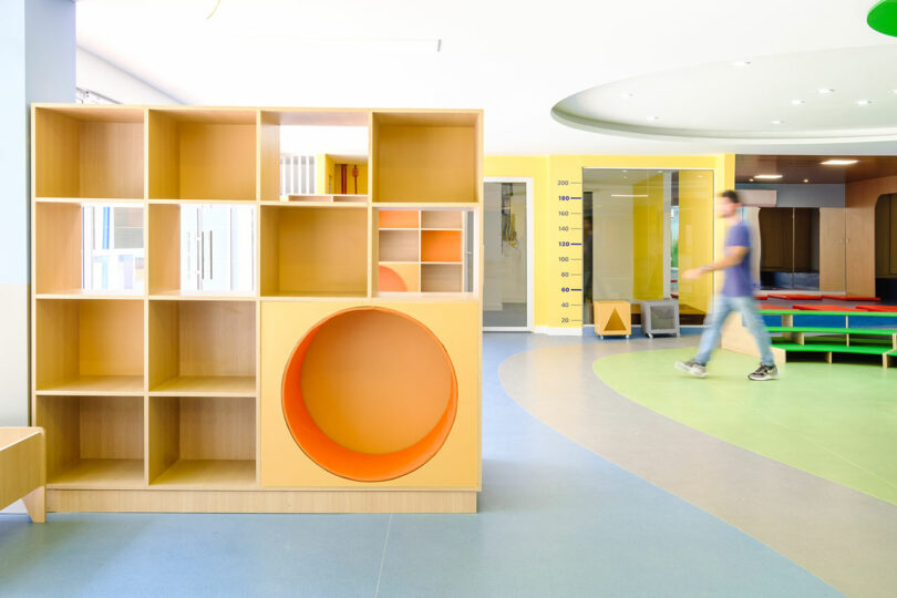 A modern, brightly lit children's play area with colorful furniture and a shelving unit featuring a large circular cutout. A person walks by, slightly blurred, in the background.