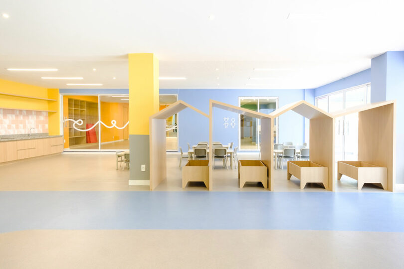 Bright, open preschool classroom with blue and yellow walls, small tables and chairs, and cubby-like wooden structures. Large windows on the right side allow natural light to fill the space.