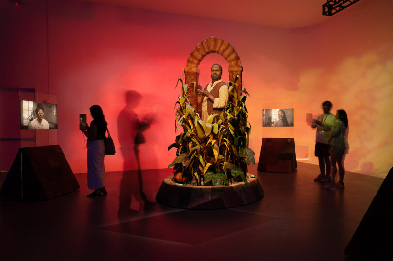 Art installation with a central statue surrounded by corn plants, set against a warm, colorful background.