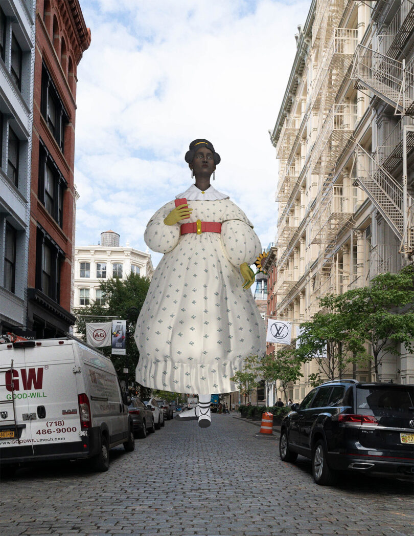 A large statue depicting a Black woman in a white dress holding a book and flowers stands in the middle of a cobblestone street lined with buildings and parked vehicles.