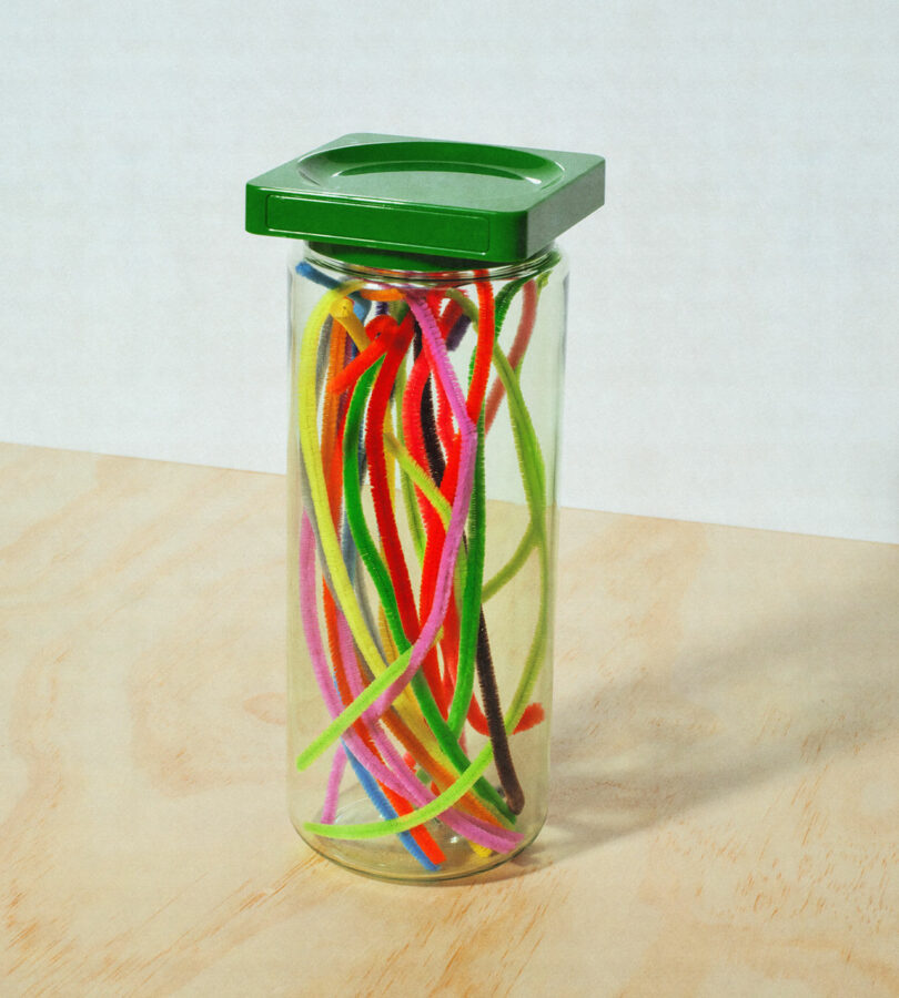 A clear jar with a green lid containing colorful pipe cleaners in various shades of red, yellow, green, blue, and pink, placed on a wooden surface.